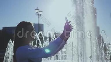 一个迷人的女孩在智能手机上自拍，站在城市的喷泉旁。 高清，1920x1080。 慢动作。